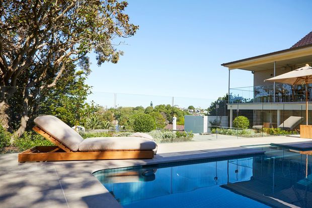 Sunny residential outdoor pool area with a blue swimming pool in the foreground, a timber-framed cushioned sun lounger, paved pool surrounds, landscaped garden, and a modern glazed house with balcony and broad roof overhang in the background.