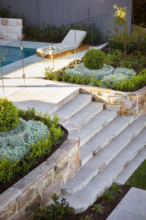 Landscaped outdoor pool area with broad pale stone steps leading to a paved terrace and swimming pool. A frameless glass safety barrier surrounds the pool, with a white reclining sun lounger on the deck. Raised stone retaining walls, clipped shrubs, ornamental grasses and layered planting create a refined garden setting.