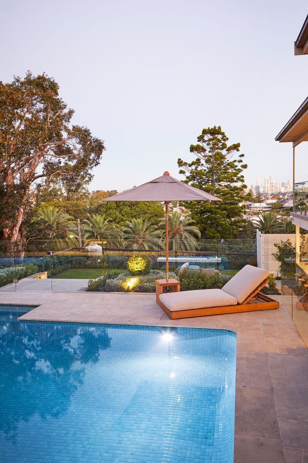 A landscaped residential pool area viewed from a terrace, featuring a curved blue-tiled swimming pool, paved pool deck, adjustable timber sun lounger, large patio umbrella, glass safety fencing, manicured garden, palms, and distant urban buildings. The scene has warm evening light and an elevated outlook.