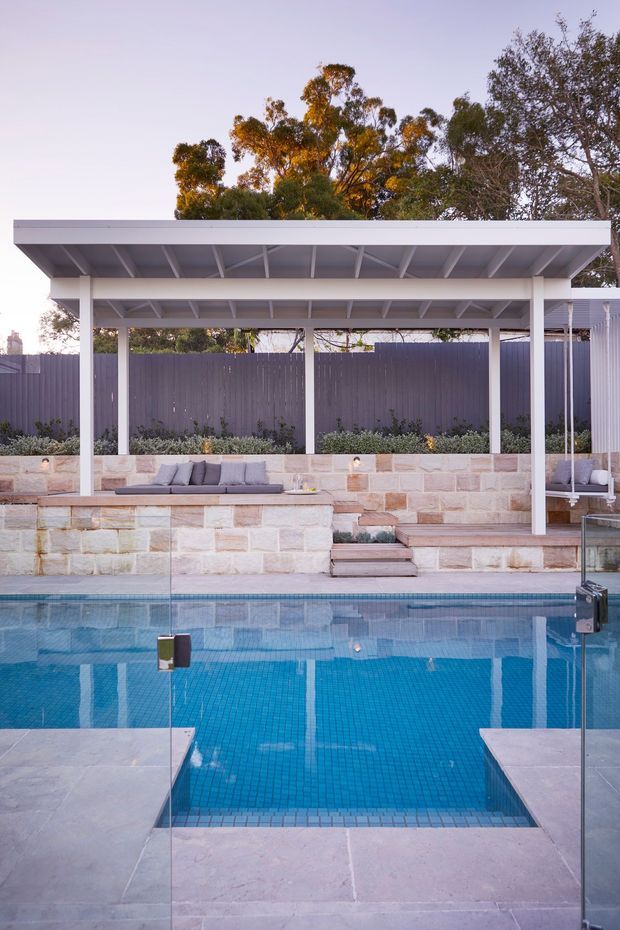 A landscaped outdoor pool area with a vivid blue tiled swimming pool in the foreground. Behind it is a raised stone-paved terrace with integrated bench seating, pale sandstone retaining walls, planting, and a white flat-roofed pergola with exposed beams. A tall dark privacy fence and mature trees form the backdrop.