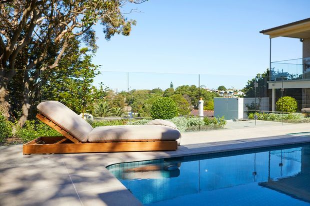 A bright outdoor swimming pool beside a contemporary home, with a timber-framed sun lounge on a paved pool deck. Glass balustrades, landscaped planting, and a broad suburban outlook are visible beyond the pool.