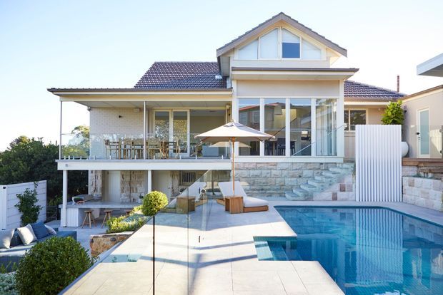 Bright exterior view of a substantial two-storey residential home with a tiled pitched roof, light rendered and stone-clad walls, large glazed openings, and a generous elevated terrace overlooking a swimming pool. The pool deck includes outdoor seating and umbrellas, with landscaped planting around the edges.