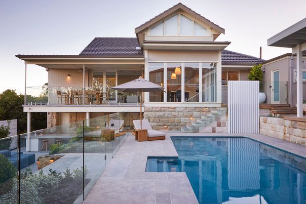 Exterior view of a substantial multi-level suburban home with a pitched gabled roof, pale rendered and masonry walls, broad covered outdoor dining terrace, extensive glazing, stone retaining walls, glass balustrades, and a swimming pool in the foreground. The design combines traditional roof and facade cues with contemporary outdoor living elements.