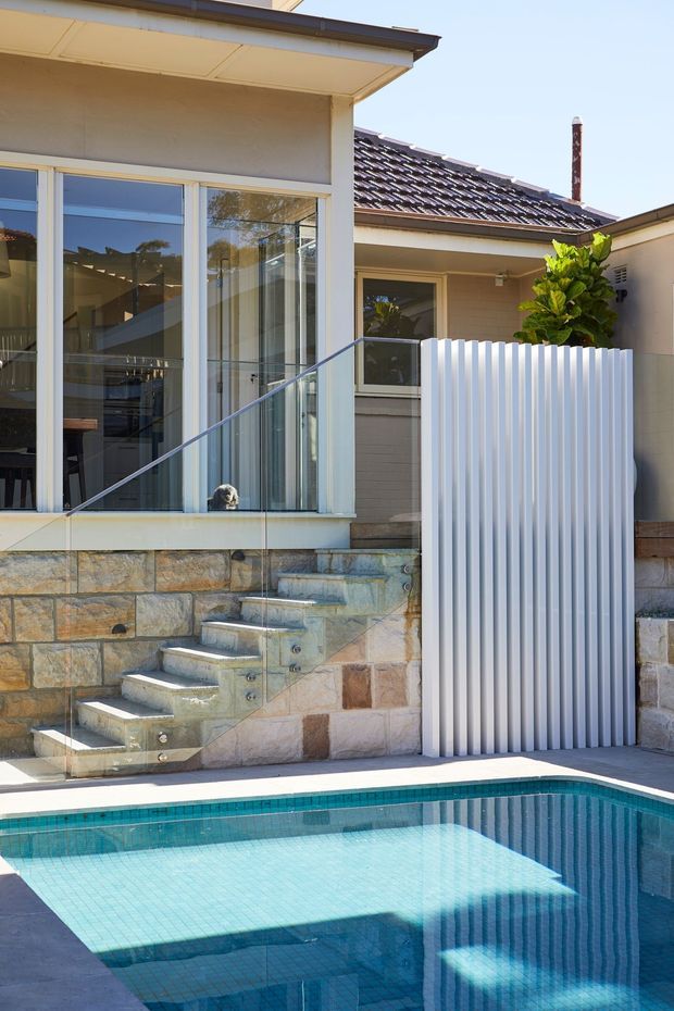 A sunny residential pool courtyard viewed from outside, featuring a turquoise tiled swimming pool in the foreground, pale stone retaining walls and steps, frameless glass balustrade, large glazed doors, a corrugated white privacy screen, and a tiled pitched roof. The architecture combines a compact contemporary addition with an older suburban dwelling.