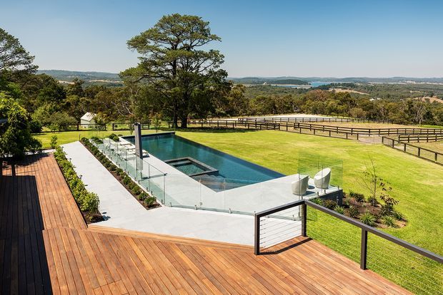A contemporary outdoor pool terrace overlooking expansive manicured lawns and fenced rural countryside. The image features a large timber deck, a rectangular in-ground swimming pool with a shallow lounging area, clear glass balustrades, pale concrete paving, outdoor seating, and landscaped planting. Rolling hills and farmland are visible in the distance.