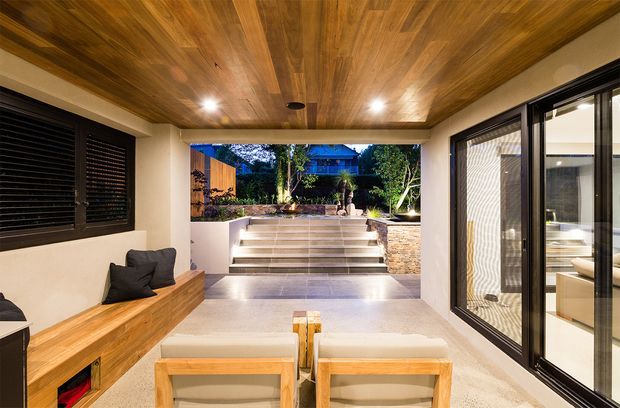 A refined covered outdoor entertaining and living area with a warm timber-lined ceiling, built-in timber bench seating, low lounge chairs, large black-framed glazed doors, and a landscaped courtyard beyond. The space opens directly onto broad steps and a planted garden, creating a strong indoor-outdoor connection.