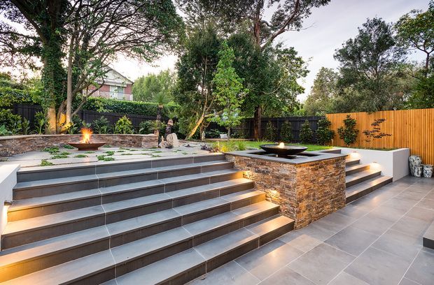 A landscaped outdoor entertaining area with broad dark tiled steps, integrated warm lighting, paved terraces, raised stone-clad fire features, planting, mature trees, and timber and dark fencing. The design creates a series of level changes and seating or gathering zones in a private garden setting.