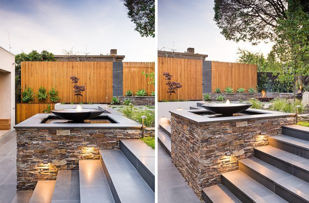A landscaped modern courtyard or garden terrace featuring a raised rectangular water feature with a dark bowl fountain, clad in stacked natural stone. Broad dark tiled steps lead around the feature, with warm integrated step and wall lighting, timber privacy fencing, planting, and a contemporary residential setting.