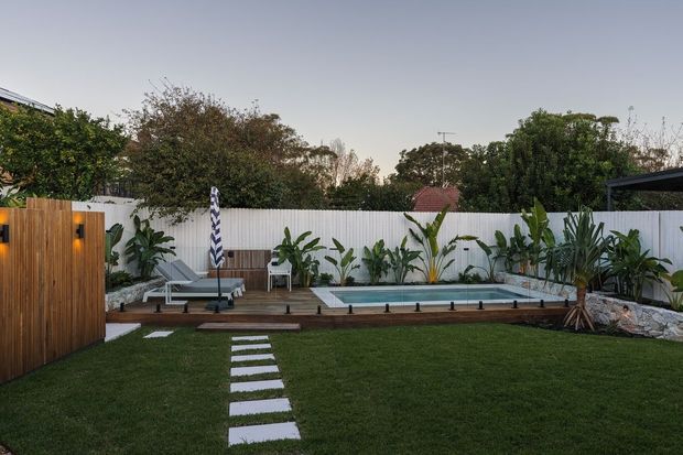 A landscaped backyard with a narrow rectangular swimming pool, white boundary fencing, tropical planting, timber screening, a paved stepping-stone path through a lawn, and poolside loungers. The scene is photographed outdoors in soft evening light.