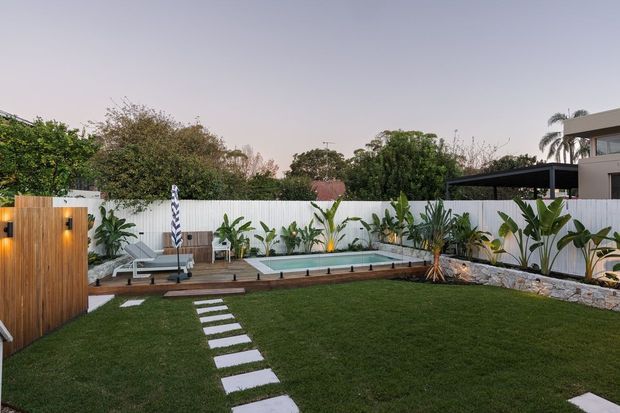 A landscaped residential backyard with a rectangular swimming pool, timber pool deck, sun loungers, tropical planting, a manicured lawn, and a stepping-stone path leading through the garden. A timber privacy screen, white boundary fence, and modern home structure frame the scene.