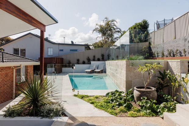 Sunlit landscaped courtyard with a rectangular swimming pool, paved terrace, timber and glass pool fencing, rendered and brick residential buildings, raised garden beds, and a large decorative planter. The space has a contemporary suburban outdoor entertaining feel with stepped levels and retaining walls.