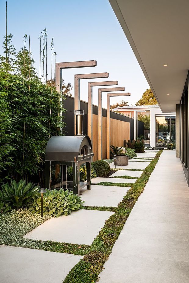 A landscaped side courtyard and walkway beside a contemporary home. Large pale concrete stepping slabs are set into low groundcover, bordered by dense bamboo and sculpted planting. A freestanding metal outdoor pizza oven sits among the garden beds, while a timber privacy fence and repeated overhead timber-and-metal frames create a strong linear rhythm. The covered building edge on the right features a smooth concrete floor, recessed downlights, and dark-framed glazing.