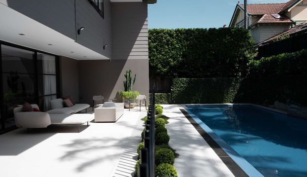 Contemporary residential courtyard with a minimalist outdoor lounge adjoining a rectangular swimming pool. The scene features clean-lined architecture, large dark-framed glazing, pale paved surfaces, sculpted hedge planting, and neighboring pitched-roof homes visible beyond the garden enclosure.