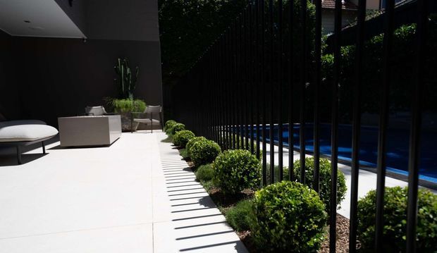A modern outdoor pool terrace with a bright white paved patio, minimalist outdoor lounge furniture, sculptural potted plants, clipped shrubs, and a black vertical-bar fence separating the terrace from the blue swimming pool. Strong sunlight creates graphic striped shadows across the paving.