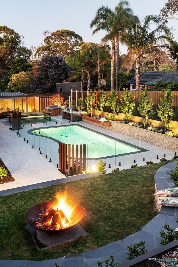 A landscaped residential backyard at dusk featuring a curved swimming pool with frameless glass safety fencing, pale paving, raised stone garden beds, timber privacy fencing, lush planting, a small covered outdoor structure, and a circular fire pit set into the lawn.