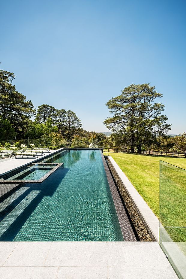 A long rectangular swimming pool with dark teal mosaic tile lining occupies the foreground, including a raised square spa or shallow water feature at one end. Pale stone paving surrounds the pool, with glass safety fencing, lounge chairs, and manicured lawn beyond. Mature trees and a distant rolling landscape sit under a clear blue sky, creating a spacious landscaped outdoor setting.