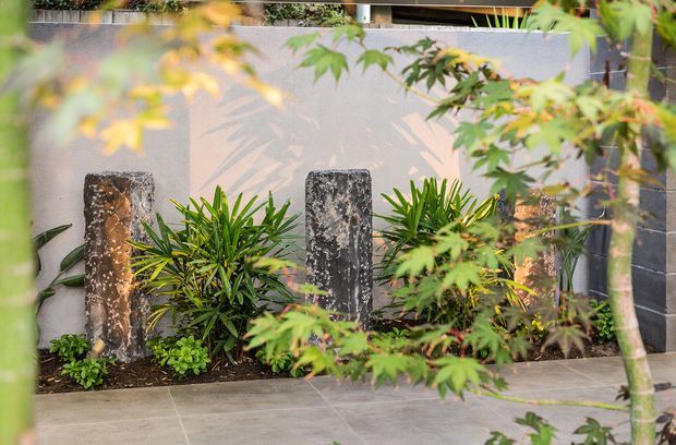A landscaped courtyard garden viewed through leafy planting, featuring a pale rendered wall with shadow patterns, dark stacked-stone wall sections, three upright natural-stone feature pillars, lush tropical-style plants, and large-format grey tiled paving.