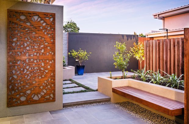A contemporary landscaped courtyard or outdoor entry area with large grey paving slabs, gravel inlays, raised rendered planter beds, timber bench seating, a warm timber fence, and a striking rust-toned perforated decorative screen set into a rendered wall. Potted and planted greenery add contrast against the neutral and warm material palette.