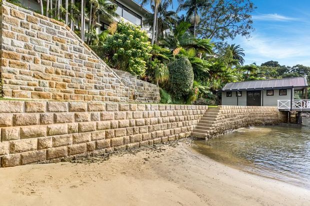 A landscaped waterfront property with extensive stepped sandstone retaining walls descending to a sandy shoreline and calm water. Dense tropical planting surrounds a dark-clad building with a low pitched metal roof and a small white-railed waterfront deck or jetty.