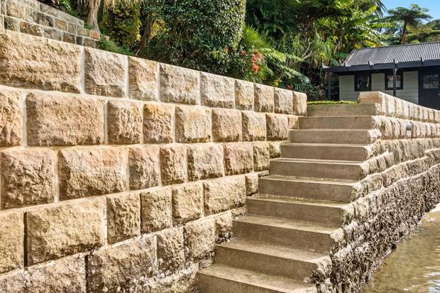 Exterior landscaped setting with broad concrete steps descending beside substantial rough-faced sandstone retaining walls. Dense tropical planting surrounds a small dark-clad building with a corrugated roof in the background; shallow water is visible at the lower right.
