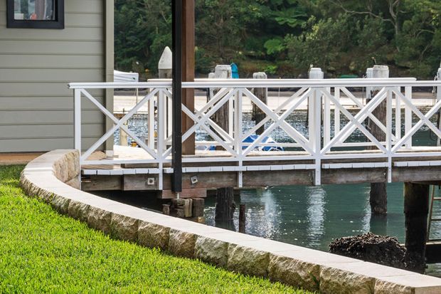 Exterior view of a waterfront deck or boardwalk beside a light horizontal-clad building. The deck features white painted timber cross-braced balustrades, dark timber posts, and a curved rough-stone retaining edge bordering maintained grass and water.