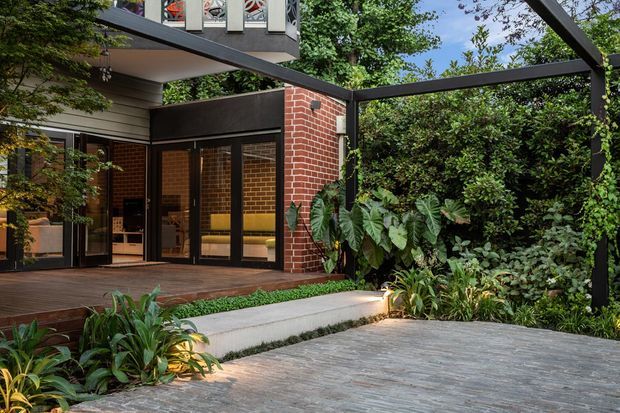 A landscaped outdoor entertaining area beside a contemporary brick-and-timber residence. The scene features a covered timber deck with large black-framed glazed doors, a red brick wall, dark structural pergola beams, concrete steps and paving, and dense tropical planting surrounding the terrace.