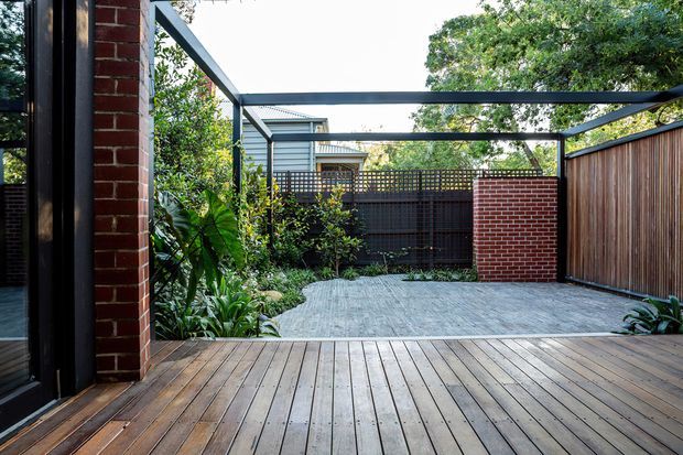 A landscaped residential courtyard viewed from a timber deck, featuring a paved outdoor area, planted garden beds, a dark steel pergola frame, brick privacy walls, timber fencing, and a partially visible dwelling beyond.