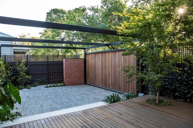 A landscaped residential outdoor courtyard or patio with a timber deck in the foreground, grey paved flooring, timber screening and fencing, a red brick wall, dark steel pergola framing, and established greenery. The space appears private and sheltered, with a small tree planted through the deck.