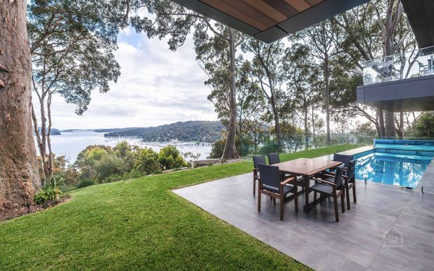 A landscaped backyard terrace overlooking a calm waterfront inlet, with a rectangular swimming pool, outdoor dining table and chairs, and a substantial timber-lined roof overhang. Mature trees frame the view, while a modern elevated balcony with glass balustrading is visible at right.