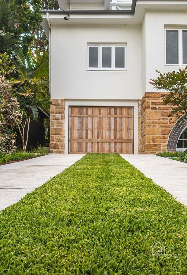 Exterior view of a residential house focused on a timber garage door, pale rendered walls, sandstone-clad lower facade, white-framed windows, and a concrete driveway divided by a narrow strip of lawn. Lush tropical landscaping borders the house.