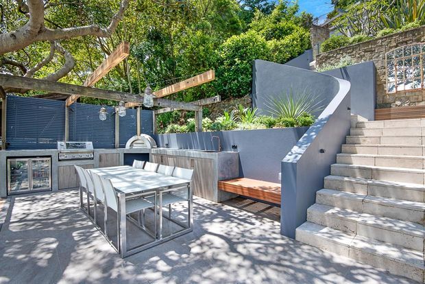 A bright landscaped outdoor entertaining area with a dining table and chairs, built-in outdoor kitchen and barbecue, timber pergola structure, bench seating, sculptural rendered walls, and broad concrete steps leading to an upper level. Mature trees and planting create a leafy, shaded setting.