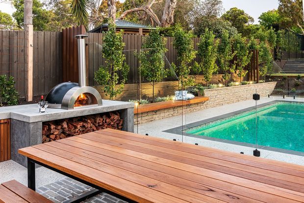 Outdoor entertaining area with a built-in pizza oven and firewood storage beside a swimming pool. A large timber dining table sits in the foreground, with rendered masonry walls, timber fencing, climbing greenery, paved surfaces and glass pool fencing.
