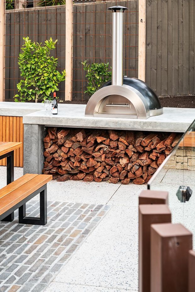 A contemporary outdoor entertaining area featuring a built-in stainless-steel wood-fired pizza oven on a concrete counter, with stacked firewood storage beneath. Timber screening, planted greenery, terrazzo-style paving, brick inlay paving, and timber seating complete the courtyard setting.