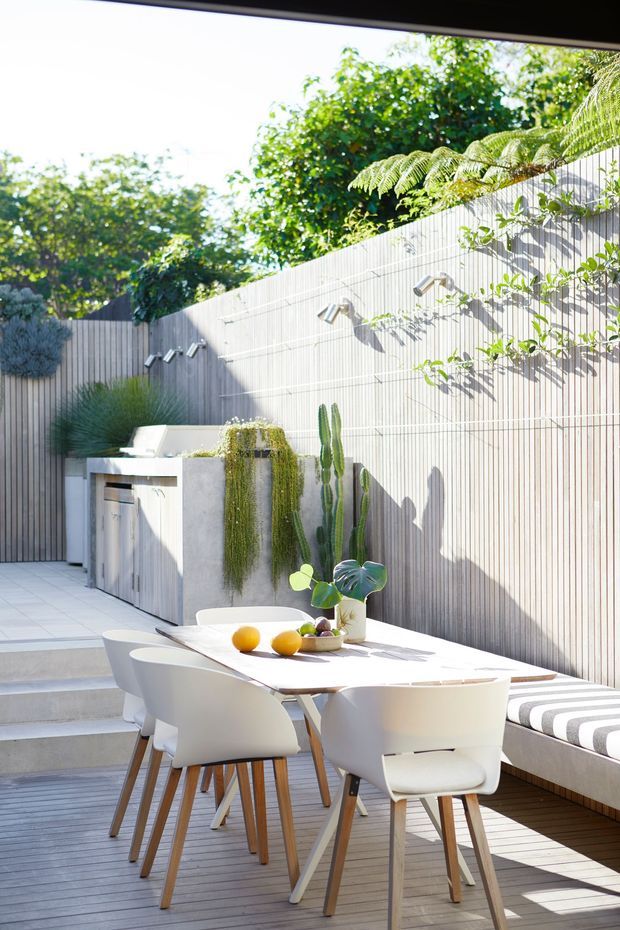 Bright contemporary outdoor dining terrace with a large light-colored dining table and white chairs on a timber deck. A built-in white upholstered bench runs along a tall pale vertical-screen fence, complemented by integrated wall lighting, raised planters, trailing greenery, sculptural cacti, and steps leading to a higher paved area.