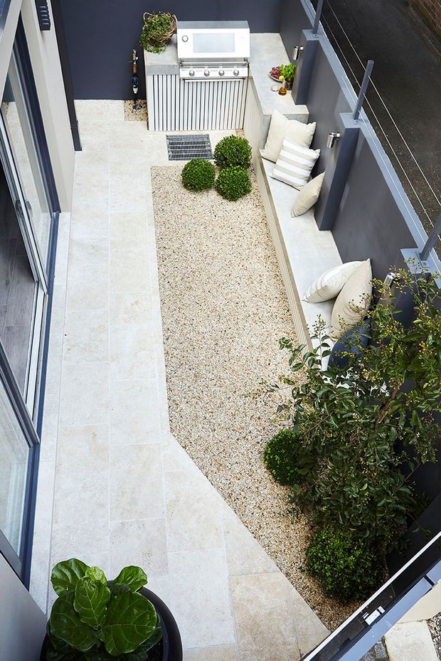 A narrow, modern courtyard viewed from above, with pale stone paving surrounding a central gravel garden bed planted with clipped shrubs and larger greenery. A built-in outdoor kitchen with a stainless-steel barbecue sits at the far end, while a long integrated bench with cream cushions lines the dark grey boundary wall. Glass and metal privacy fencing frames the space.