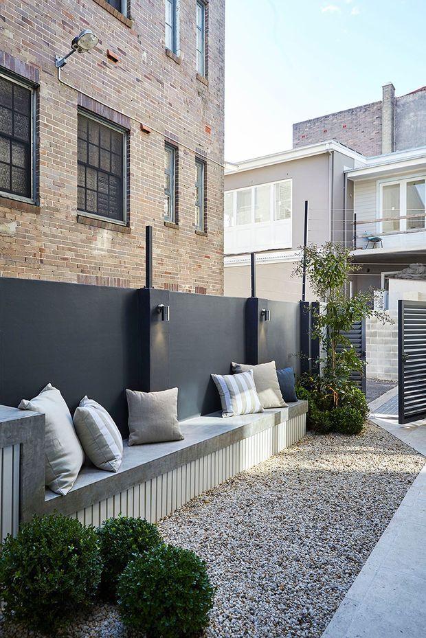 A compact landscaped courtyard between urban brick buildings, featuring a long built-in concrete bench with vertical ribbed cladding, pale cushions, a tall dark privacy wall, gravel paving, clipped shrubs, and a young tree. Contemporary residential architecture surrounds the courtyard, with exposed brick facades, metal-framed windows, and a small balcony.