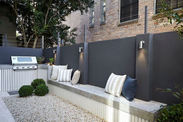 A compact urban outdoor entertaining area with a built-in stainless-steel barbecue, a long fixed concrete bench with a fluted white base, striped and neutral cushions, dark rendered privacy walls, and a gravel-covered courtyard. A brick building facade and integrated wall lighting form the backdrop.