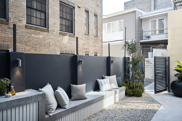 A contemporary urban courtyard with a long built-in concrete bench and pale cushions along a dark charcoal privacy wall. The courtyard has a light gravel surface, vertical ribbed detailing beneath the bench, exposed light brickwork, modern multi-storey surrounding buildings, black-framed windows, planting in large pots, and a louvred metal gate.
