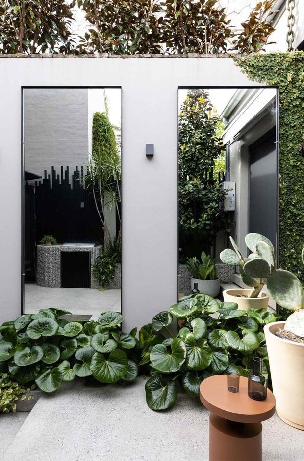 A compact contemporary courtyard viewed from within, featuring white rendered walls, large black-framed mirrors, abundant glossy green planting, pale terrazzo or concrete paving, and a small sculptural round outdoor table. The mirrors visually extend the planted courtyard and reflect a dark decorative screen, planters, and surrounding architecture.