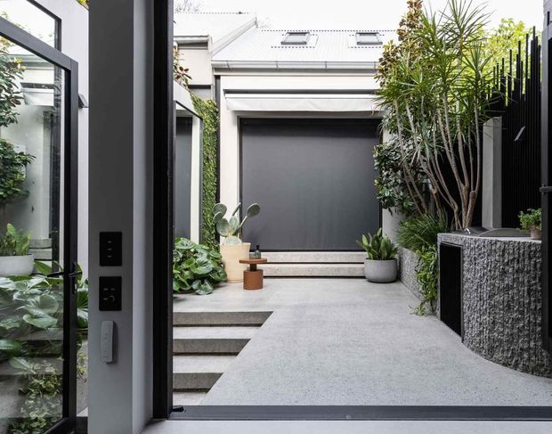 A contemporary courtyard entry viewed from inside through an open glazed door. The compact paved space features a charcoal garage or entry door beneath a light metal roof with skylights, layered concrete steps, dense potted greenery, a textured dark stone-clad planter or wall, and black architectural framing. The scene is bright, refined, and modern with a minimalist palette.