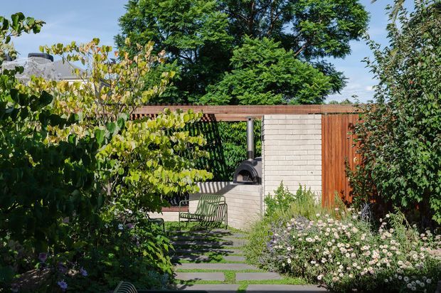 A lush landscaped courtyard or garden with a stepping-stone pathway leading to a compact outdoor entertaining area. The space features a pale brick masonry wall, warm vertical timber fencing, a freestanding dark outdoor fireplace or pizza oven, and a metal garden chair, surrounded by mature trees and flowering planting.