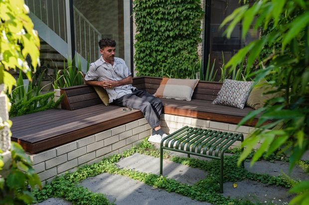 A leafy outdoor courtyard or terrace beside a modern building, featuring an L-shaped built-in timber bench with upholstered cushions, raised light brick planter walls, lush vertical greenery, and a green slatted metal coffee table. A person is seated on the bench, creating a relaxed garden lounge setting.