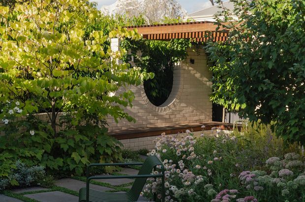 A lush landscaped courtyard or garden seating area framed by mature trees and flowering plants. A light brick wall with a distinctive oval opening sits beneath a timber pergola or slatted roof structure, with a dark green outdoor chair in the foreground and partial mountain scenery visible beyond.