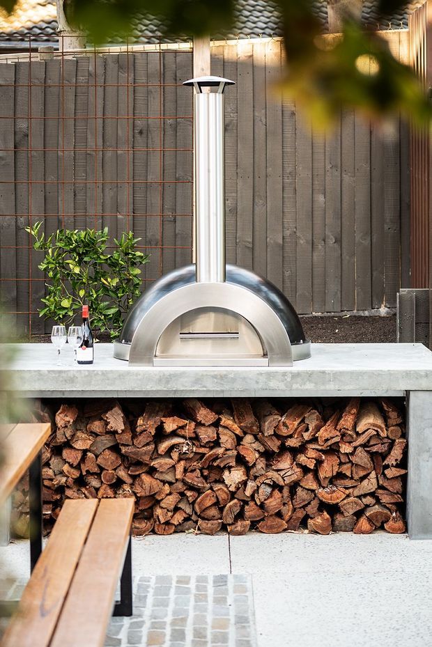 A compact outdoor entertaining area featuring a built-in wood-fired pizza oven with a tall stainless-steel flue, set on a concrete benchtop above an open wood-storage niche. The oven is backed by a dark vertical timber privacy fence with a red metal trellis, while blurred greenery frames the scene. Timber decking and light tiled paving complete the courtyard setting.