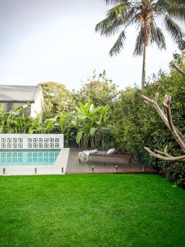 A landscaped backyard with a rectangular swimming pool, patterned privacy wall, timber pool deck, sun lounger, lush tropical planting, palm trees, and a broad green lawn. A small light-coloured house with a corrugated metal roof is partially visible behind the garden.