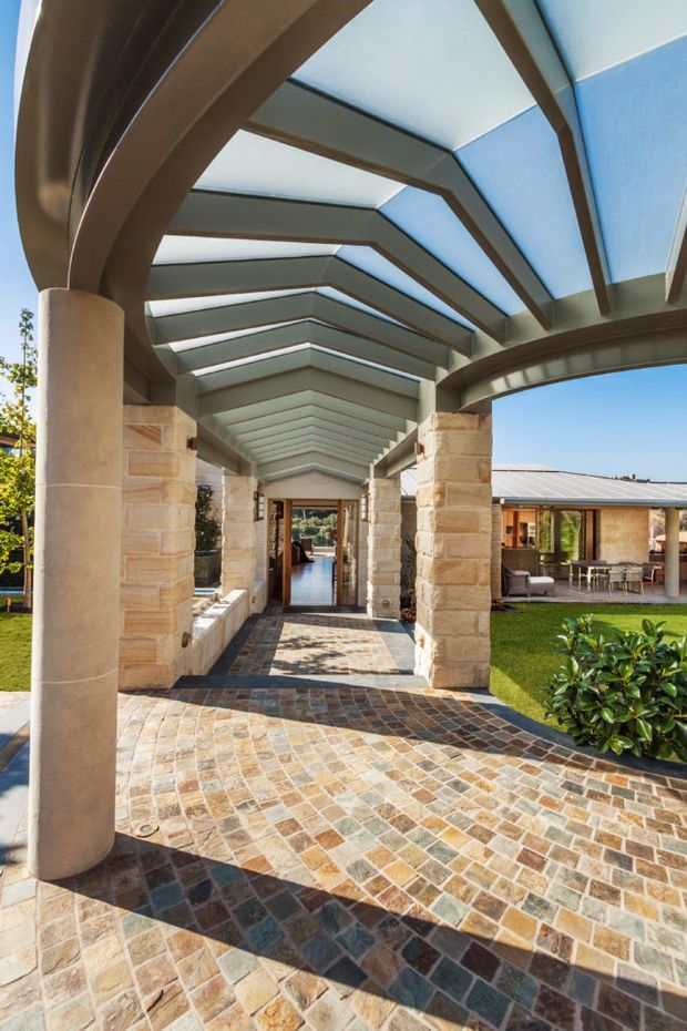 Covered architectural walkway or entry arcade connecting sections of a stone-clad residence. The space features substantial curved and straight structural beams, stone columns, translucent roof panels with abundant daylight, and a multicoloured patterned stone or tile paving surface. A glazed doorway and living areas are visible at the end, with landscaped lawn and garden visible to the sides.