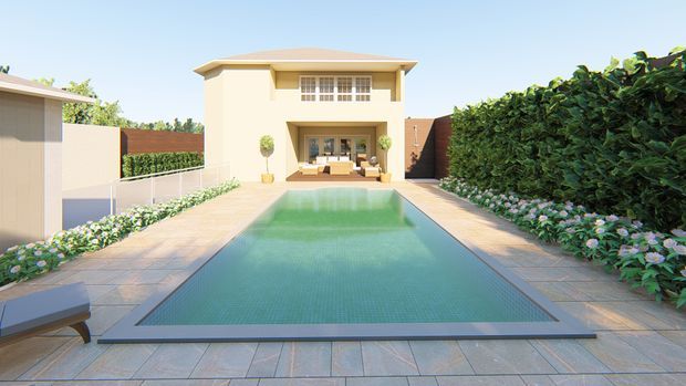 A landscaped backyard with a long rectangular swimming pool leading toward a two-storey cream-coloured house. The pool is surrounded by pale paved terraces, clipped hedges, flowering borders, and a small outdoor seating area beneath a deep upper-level opening. The scene has a clean, refined residential courtyard feel in bright daylight.