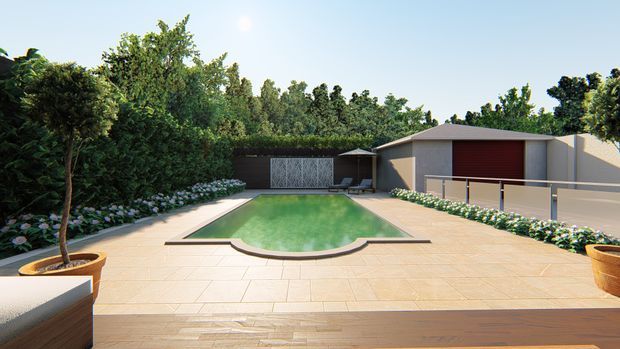 A landscaped outdoor pool courtyard viewed from a timber deck. The scene features a pale tiled pool terrace, green swimming pool with a curved end, lush planted borders, potted trees, and a detached single-storey outbuilding with a broad hipped roof and red garage door. A privacy fence and gate sit behind the pool.