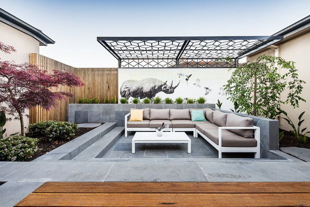 A contemporary landscaped courtyard or outdoor entertaining area with a sunken grey-paved lounge zone, a large L-shaped outdoor sectional, white coffee table, raised concrete planter and seating walls, timber privacy fencing, and a geometric overhead pergola. A decorative mural and established planting add visual interest, while timber decking occupies the foreground.