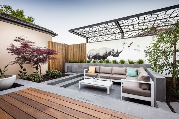 A contemporary landscaped courtyard entertaining area with a timber deck, built-in concrete seating, a large sectional sofa, white coffee table, raised garden beds, a sculptural water feature, timber screening, and a striking geometric pergola canopy. A painted mural forms a feature wall behind the seating.